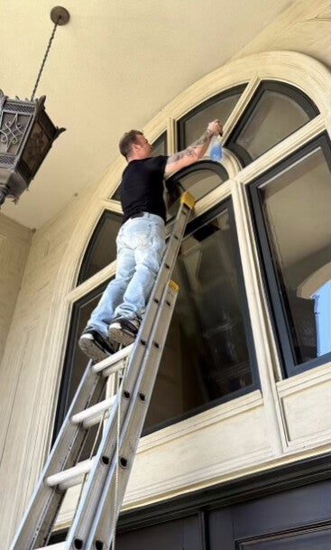A Klein's cleaner on a tall ladder cleaning the top of a large arched window from inside a two-storey foyer