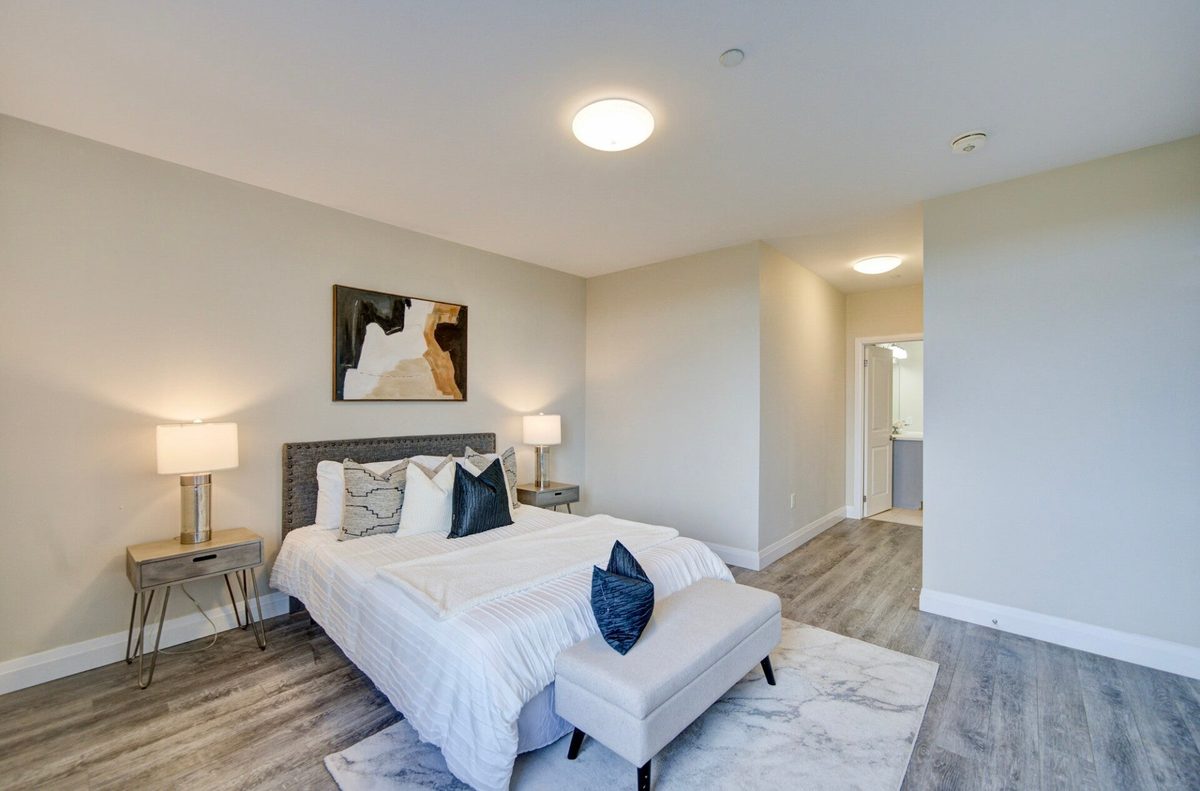 A cleaned bedroom with a made white bed, navy cushions, a bench at the foot and matching bedside lamps