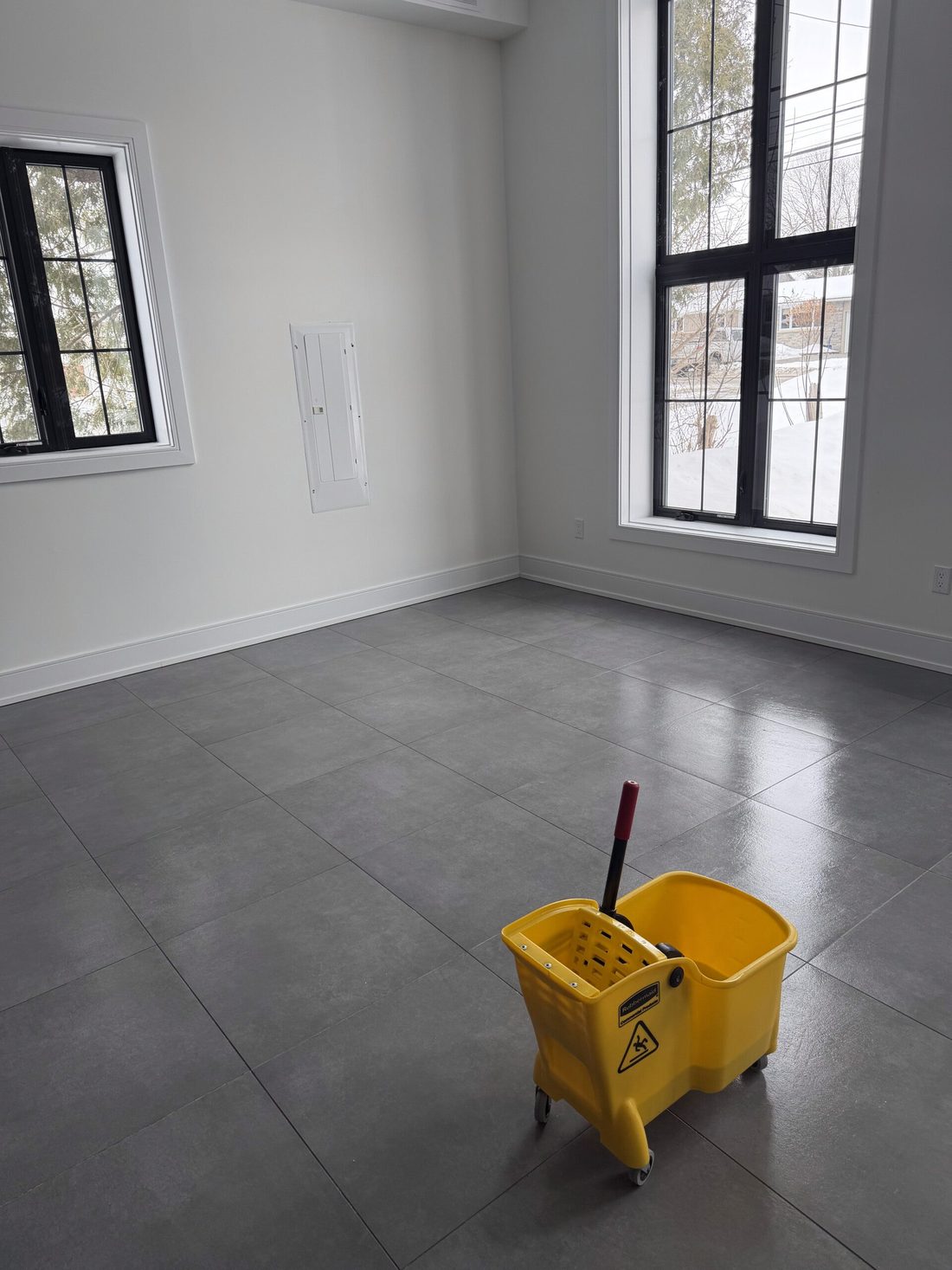 A freshly mopped grey tile floor in an empty commercial room, with a yellow mop bucket by the window