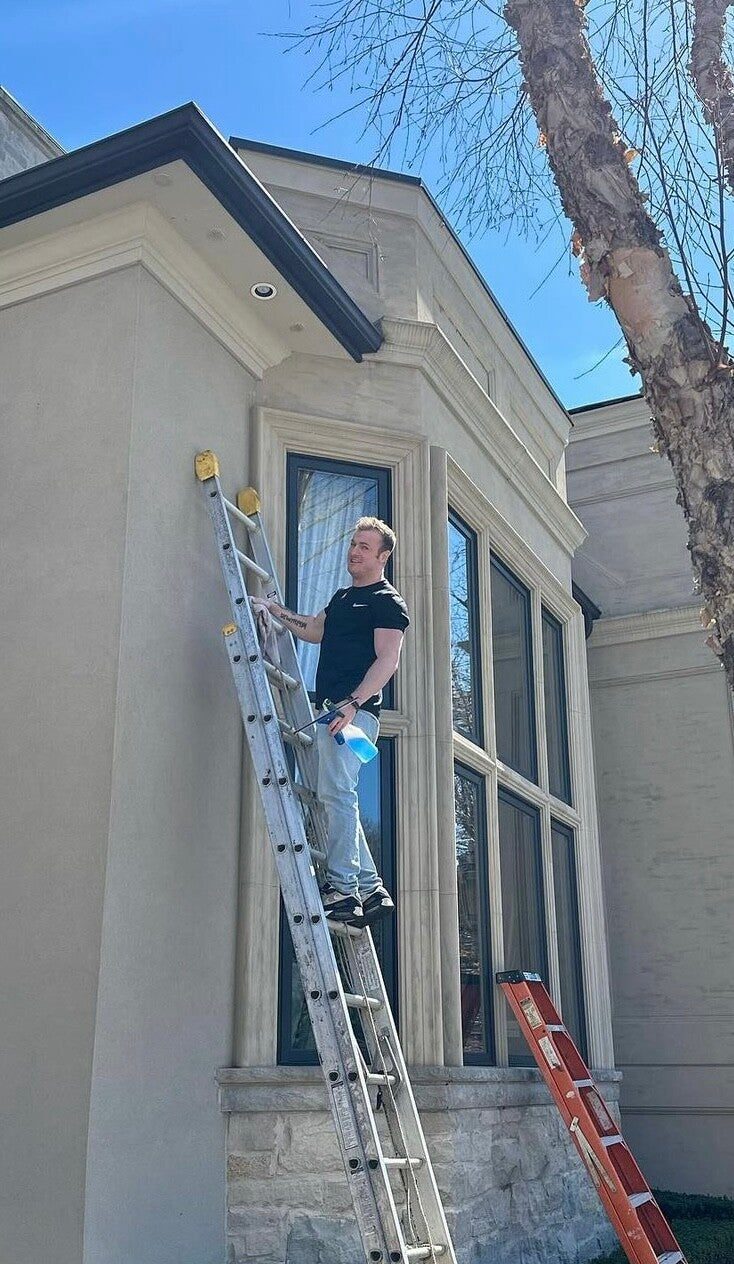 A Klein's cleaner on a ladder washing the tall front windows of a stone two-storey house on a sunny day